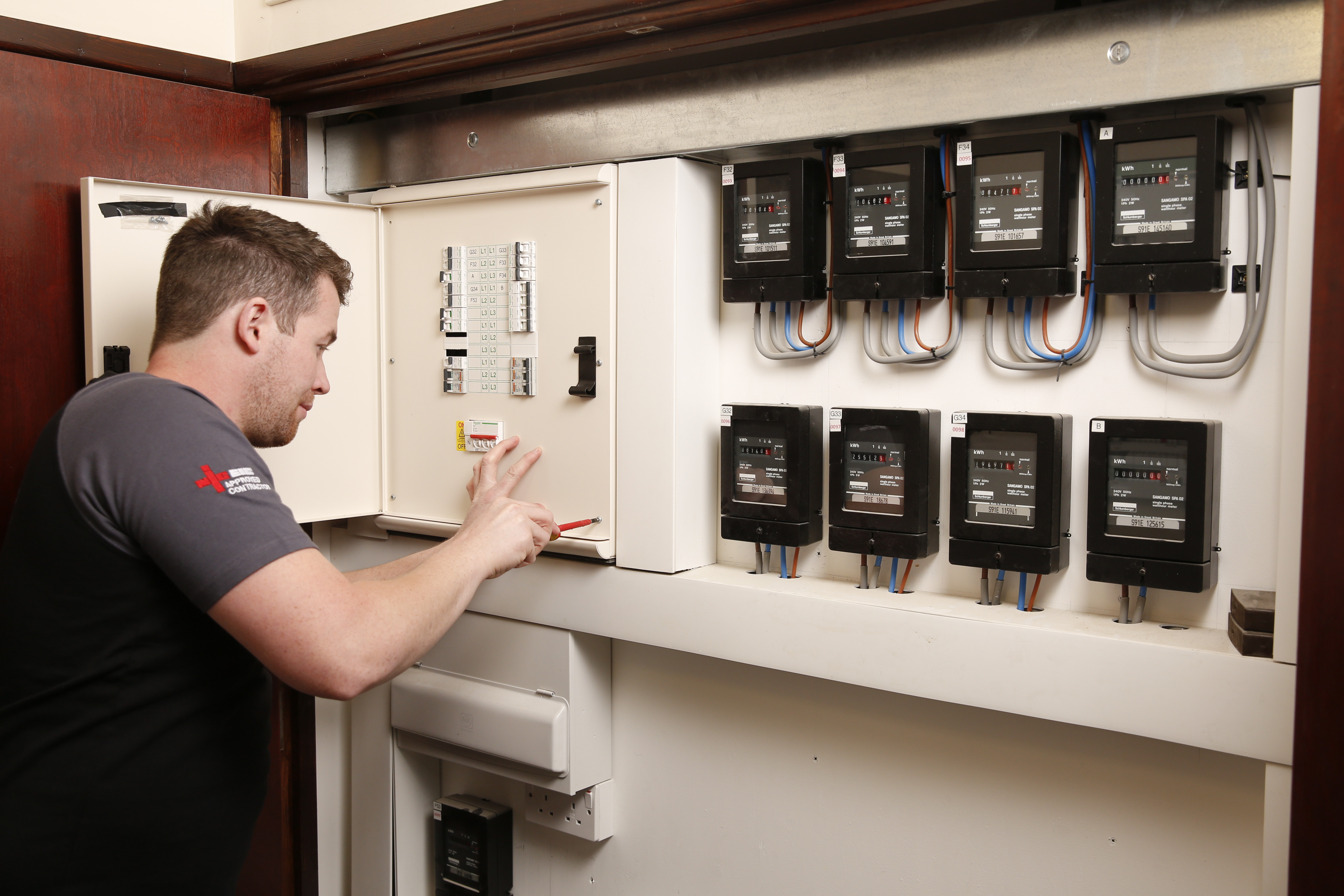 AC1Electrical worker working on a fuse box panel next to multiple electricity meters in a commercial or residential setting.